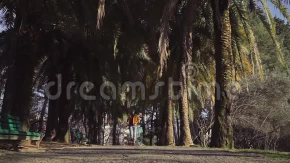 穿着牛仔裤和黄色夹克的快乐女孩带着棕榈树沿着小巷跑有时跳起来抓住他们视频的预览图