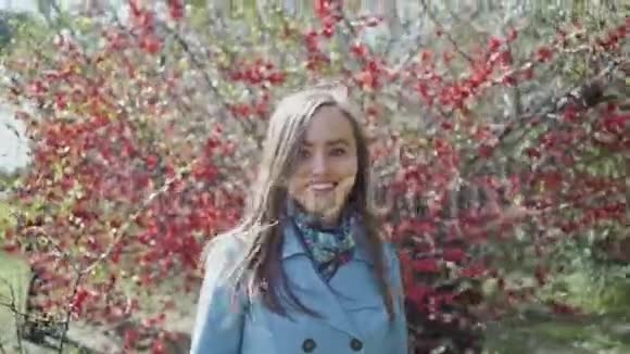穿着蓝色雨衣的快乐女孩穿过春天的公园走过一棵开着红花的树视频的预览图