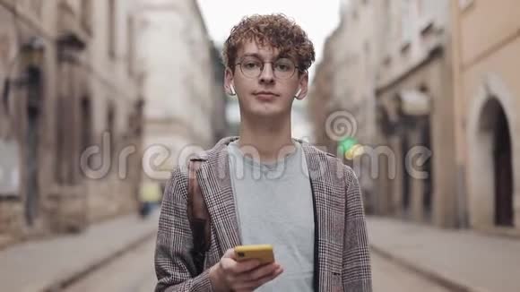 关闭冷静的千禧一代戴着耳机和眼镜看着相机手里拿着智能手机视频的预览图