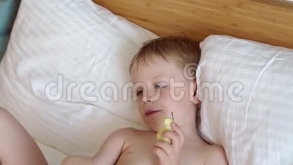 一个金发小男孩躺在一张白色的床上吃着一个苹果慢动作视频的预览图