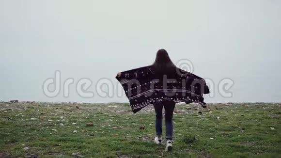 一个穿着针织斗篷的女孩在海边的悬崖边上伸直双臂奔跑视频的预览图