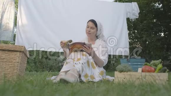 可爱的女人头上戴着一条白色披肩坐在草地上坐在晾衣绳前看书视频的预览图