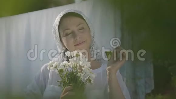 可爱的女人头上戴着白色披肩嗅着雏菊看着户外晾衣绳附近的相机洗衣日视频的预览图