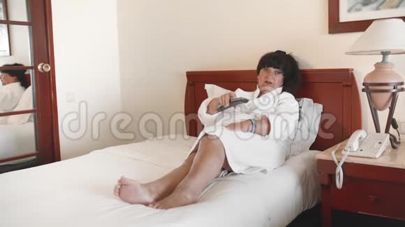 老年妇女躺在酒店的床上穿着白色浴袍用遥控器控制电视使用的概念视频的预览图