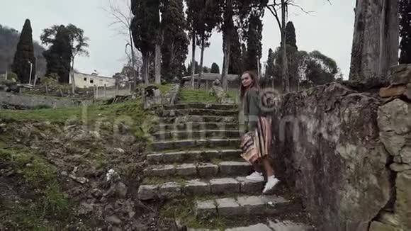 穿着条纹裙子的女孩爬上长满苔藓的岩石楼梯爬在柏树的背景下视频的预览图