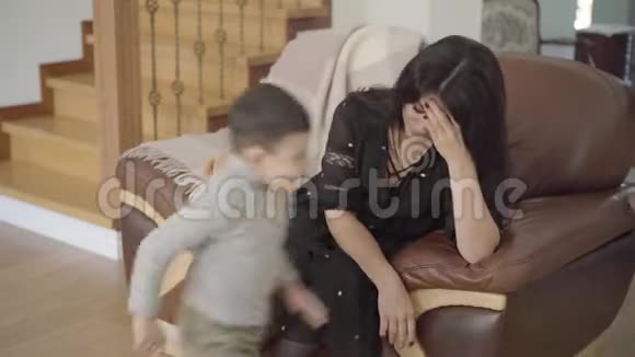 美丽的中东母亲偏头痛坐在扶手椅上她的两个小孩子跑来跑去累了视频的预览图