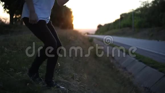 年轻人带着黑色的背包和帽子走后坐在草地上在柏油路附近日落的背景视频的预览图