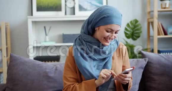 美丽的中东女人戴着头巾在家里用智能手机微笑视频的预览图