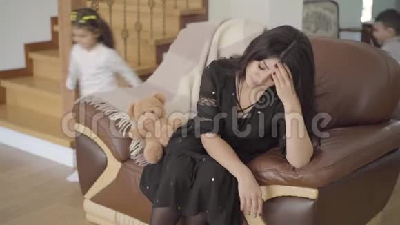 疲惫的中东母亲坐在扶手椅上她的两个孩子跑来跑去美丽的女人视频的预览图