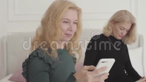 一个成熟的高加索女人向她的朋友展示智能手机的肖像富有的金发贵妇坐在家里视频的预览图