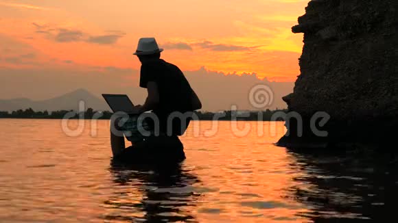在日落的背景下一个带笔记本的程序员坐在海边的岩石上从事自然研究的自由职业者遥远的视频的预览图