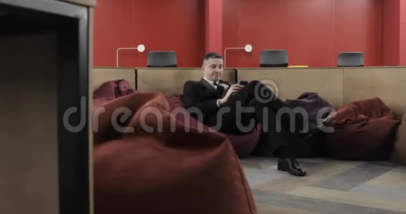 成熟男经理坐在豆袋椅上浏览办公室手机视频的预览图
