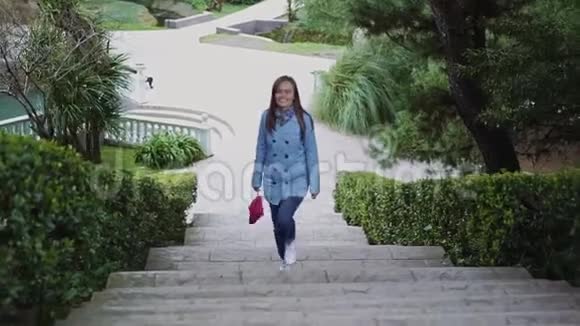 穿着蓝色外套和粉红色离合器的快乐女孩爬上公园绿色中间的白石楼梯视频的预览图