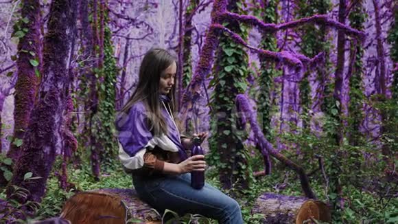 紫罗兰童话森林穿着运动夹克的女孩正坐在一片迷人的紫色森林里的树桩上拧开盖子视频的预览图