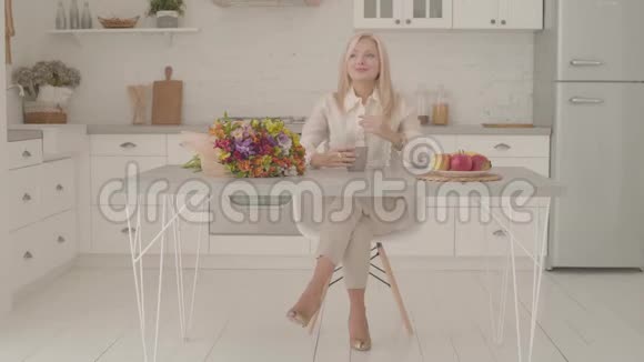 一位金发白种人女士坐在桌边端着一杯茶或咖啡和一束花快乐的成年女人视频的预览图