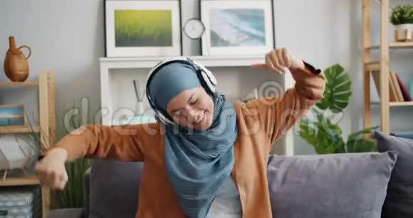 戴头巾的穆斯林女孩慢动作在家跳舞时戴耳机欣赏音乐视频的预览图