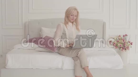 自信的金发白种人女商人坐在家里的床上在笔记本电脑上打字年轻漂亮的女士视频的预览图
