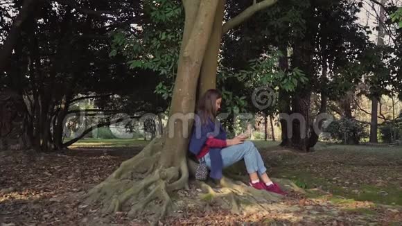 一个穿着蓝色毛衣带着智能手机的女孩正坐在一棵大树下树根缠绕得很厉害视频的预览图