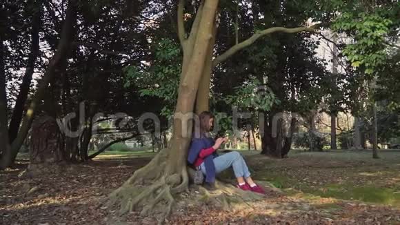 可爱的女孩手里拿着电话坐在大树下外面长出了扭曲的根视频的预览图
