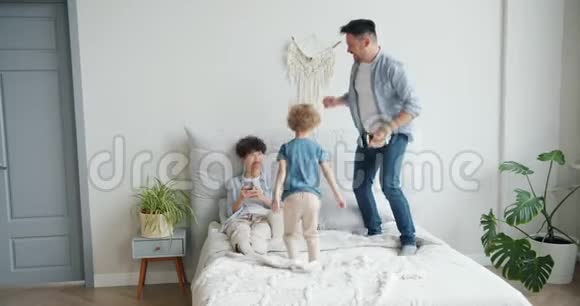 父亲和儿子在家里跳着床而母亲则用智能手机微笑视频的预览图