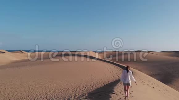 电影拍摄一个穿白色衣服的女孩沿着沙漠中的沙丘的边缘行走在大西洋的海岸视频的预览图