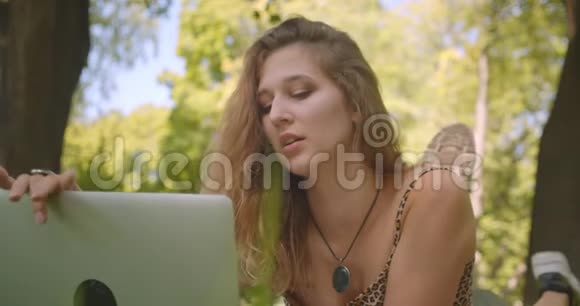一位年轻漂亮的白种人女性的特写照片她在笔记本电脑上打着视频开心地躺在草地上的地毯上视频的预览图