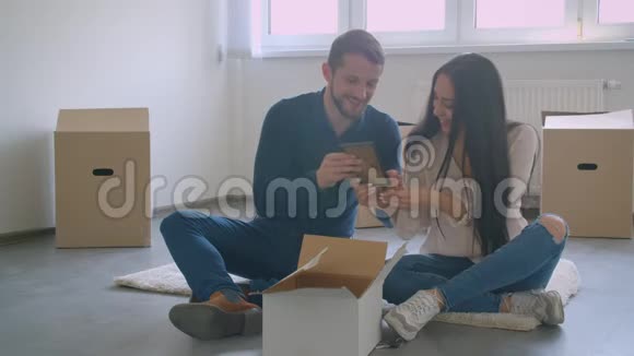 年轻的白种人夫妇坐在新房子的地板上选择了相框照片的位置视频的预览图