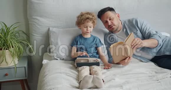 爱爸爸给孩子看书而孩子在公寓里用平板电脑视频的预览图
