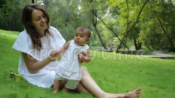 年轻漂亮的母亲穿着一件白色的衣服教她刚出生的女儿在公园里的一片新鲜的绿草上行走年轻快乐视频的预览图