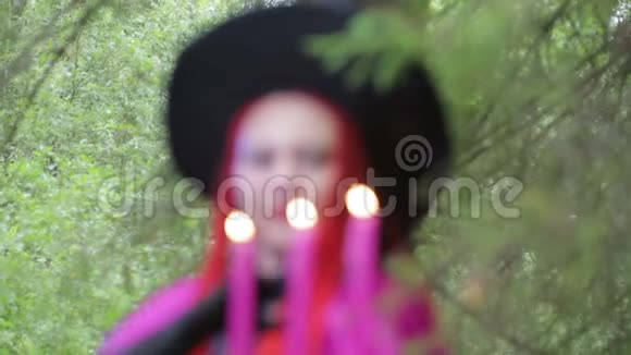 一个年轻的女巫红头发尖尖的帽子和一件覆盆子斗篷在森林里用蜡烛变魔术视频的预览图