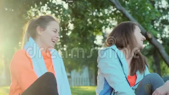 两个女孩在清晨慢跑和大笑后坐在草地上一条毛巾挂在她们的脖子上视频的预览图