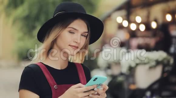 戴着黑色帽子戴着智能手机的金发女孩站在城市街道上的画像视频的预览图