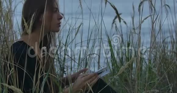 美丽的白种人黑发女孩坐在湖岸上带着书看着远方一个美丽的女人视频的预览图