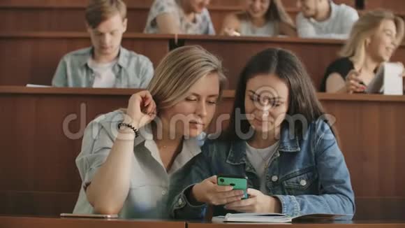 在大学演讲的休息时间手持智能手机的女学生在观众中大笑视频的预览图