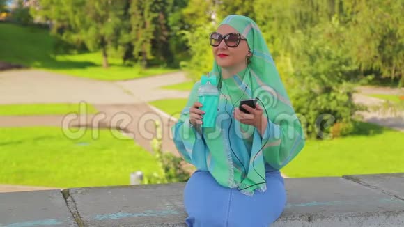 公园里一位年轻的穆斯林妇女戴着绿色围巾和太阳镜喝着咖啡正在送信视频的预览图