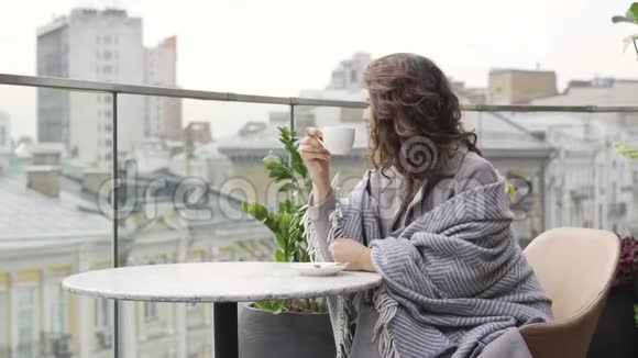 年轻的白种人女人被覆盖在毯子上喝茶或咖啡坐在露台上的桌子上她看上去视频的预览图