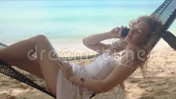 在沙滩上躺在吊床上在智能手机上说话的快乐女人视频的预览图