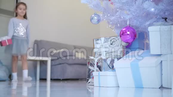 美丽的小天使女孩带着完美的微笑把一个红色的礼物盒和白色的蝴蝶结放在圣诞树下视频的预览图