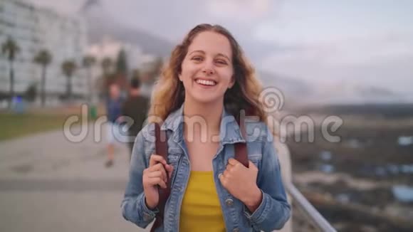 一位微笑的金发年轻女子站在城市街道上背包看着摄像机的画像视频的预览图