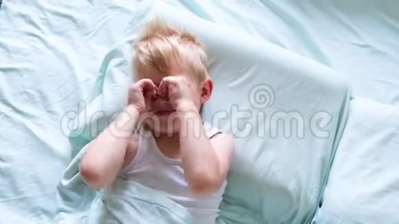 一个金发小男孩躺在床上微笑这个男孩在清晨用双手揉眼睛是时候醒来了视频的预览图