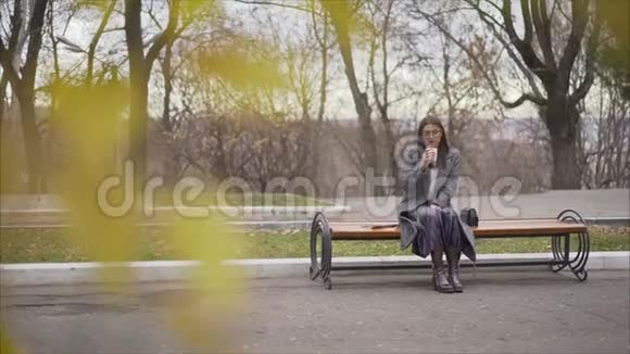 秋天在一个阳光明媚的公园里穿着外套的漂亮女孩坐在长凳上女孩喝温咖啡和户外肖像视频的预览图