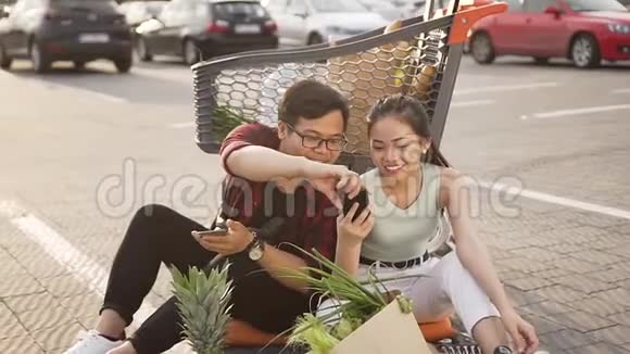 一对年轻迷人的亚洲情侣坐在市场手推车旁的地上使用智能手机视频的预览图