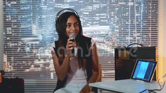 戴着耳机的漂亮黑人女孩对着麦克风唱歌在家庭录音室里录制一首歌视频的预览图