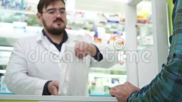 微笑的胡须药剂师把白色包裹和药物给顾客穿白色长袍的专业工人视频的预览图