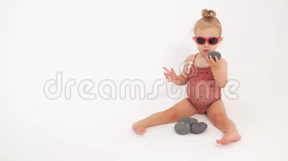 穿着泳衣和太阳镜的可爱女婴在白色背景下玩鹅卵石视频的预览图