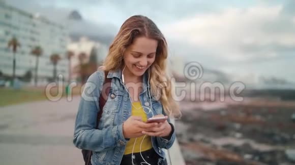 在城市街道上戴着耳机听音乐用手机浏览的金发年轻女子微笑视频的预览图
