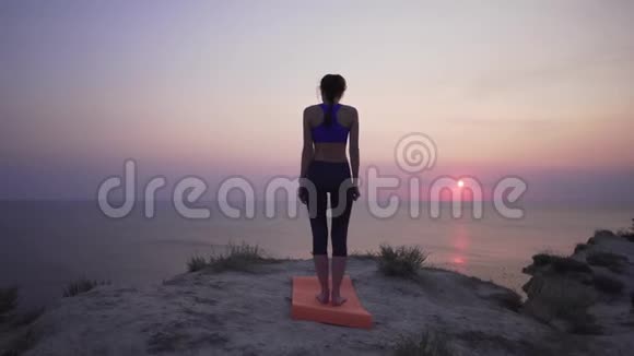 一个年轻的女人站在悬崖附近的山上做瑜伽姿势可以看到海洋和日落或黎明和平与和平视频的预览图
