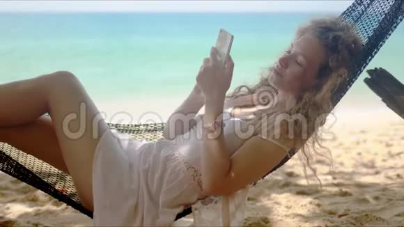 在海滩上躺在吊床上放松的女人浏览智能手机视频的预览图