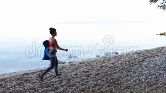 穿运动服的运动女性拎着瑜伽垫在卵石海滩散步视频的预览图