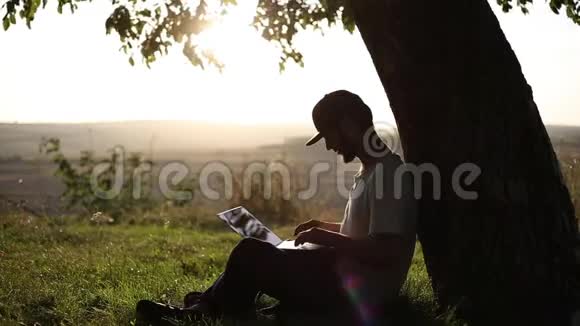 年轻人坐在公园的草地上靠在大树上腿上有笔记本电脑打字工作视频的预览图